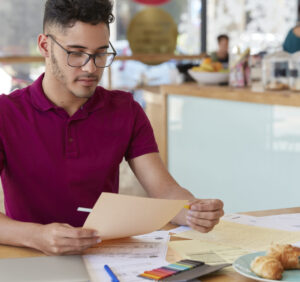 Shot of unshaven guy examines paperwork, uses stickers, dressed in casual t shirt and spectacles. Creative male blogger works with documentation, has hard working day, develops new strategy.