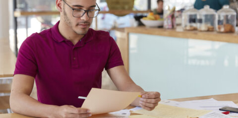 Shot of unshaven guy examines paperwork, uses stickers, dressed in casual t shirt and spectacles. Creative male blogger works with documentation, has hard working day, develops new strategy.