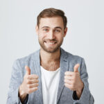 Portrait of stylish bearded man or customer with broad smile, looking at the camera with happy expression, showing thumbs-up with both hands, achieving professional goals. Body language